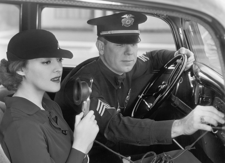 Woman sitting next to a policeman in his car holding a microphone